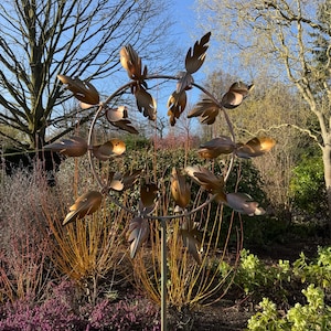 May include: A bronze metal garden sculpture with a circular design and leaf motifs. The artwork is mounted on a metal pole and set in a garden with purple and yellow plants, creating a natural aesthetic.