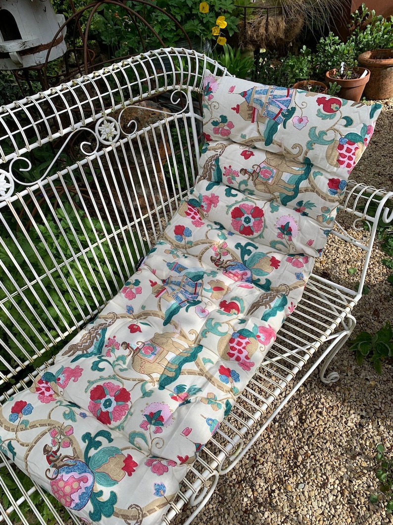 May include: A white metal garden bench with a floral patterned cushion. The cushion has a cream background with pink, blue, and green floral designs. The cushion is quilted and has a tufted design.