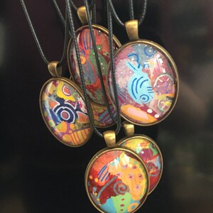 Peut inclure: Cinq pendentifs ronds colorés avec des motifs abstraits sur un fond noir. Chaque pendentif est attaché à un cordon noir.
