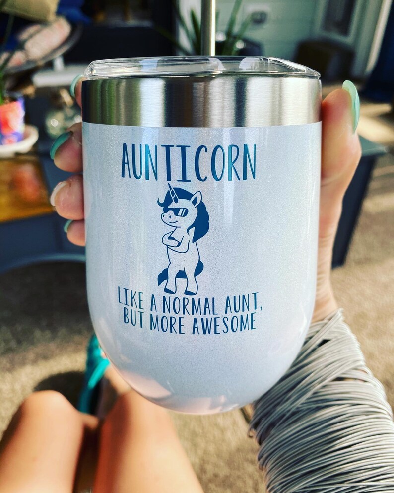 May include: A stainless steel tumbler with a white, glittery finish. It features a blue unicorn graphic with the text "Aunticorn, like a normal aunt, but more awesome".