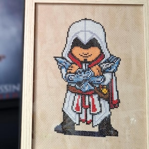 May include: A framed cross-stitch of a character from the Assassin's Creed video game series, in a wooden frame. A black and white PlayStation 5 controller is in the foreground, with a framed Assassin's Creed movie poster in the background.