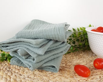 May include: A stack of folded, light teal-colored cloth napkins. The napkins are on a round, woven placemat. A white bowl with red tomatoes and green sprigs are in the background.