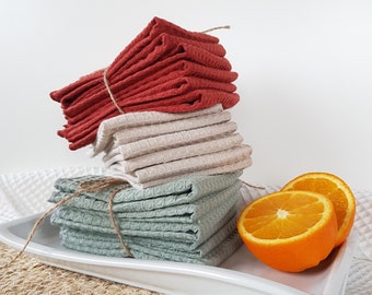 May include: A stack of three different colored waffle weave kitchen towels. The towels are folded and tied with twine. The towels are on a white plate with two orange slices on the plate. The plate is on a woven straw mat.