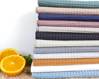 May include: A stack of waffle weave kitchen towels in various colors, including teal, beige, navy, pink, gray, white, and light blue. The towels are folded and stacked on a woven surface.