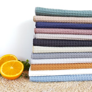 May include: A stack of waffle weave kitchen towels in various colors, including teal, beige, navy, pink, gray, white, and light blue. The towels are folded and stacked on a woven surface.