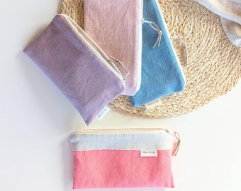 May include: Four small zippered pouches made of linen fabric in different colors: pink, purple, blue, and red. The pouches are arranged on a woven straw mat. Each pouch has a white label with the text "Flora & Lin" printed on it.