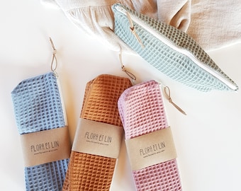 May include: Three waffle-textured fabric pouches with zipper closures. The pouches are in blue, orange, and pink. Each pouch has a label with the text "FLORA ET LIN" and a website address.