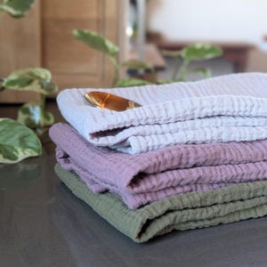 Peut inclure: Une pile de quatre serviettes en coton texturé dans les tons lavande, mauve et vert sauge. Une cuillère dorée repose sur le dessus de la pile. Les serviettes sont pliées avec soin, suggérant un thème de décoration intérieure ou de textile de cuisine.