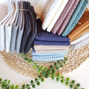 May include: A stack of waffle weave kitchen towels in various colors, including gray, black, blue, brown, and green. The towels are folded and stacked on a woven mat with green leafy plants in the foreground. The text "FLORA ET LIN" is visible on the top towel.