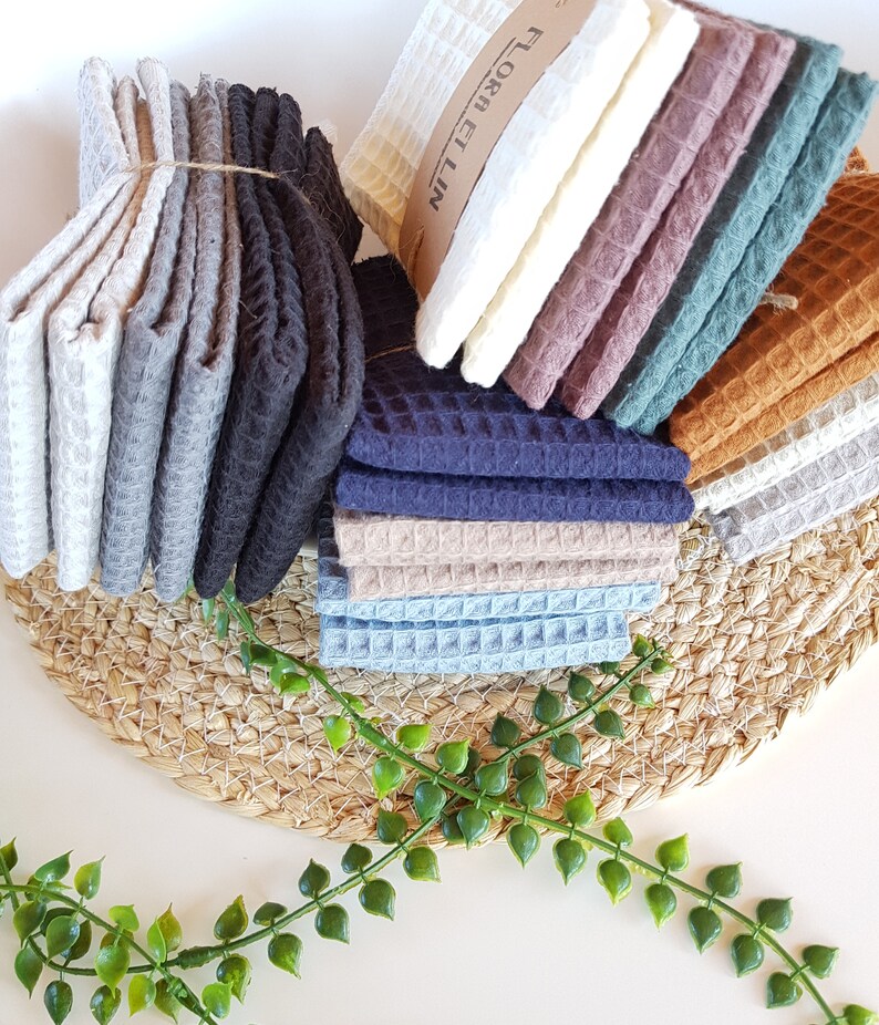 May include: A stack of waffle weave kitchen towels in various colors, including gray, black, blue, brown, and green. The towels are folded and stacked on a woven mat with green leafy plants in the foreground. The text "FLORA ET LIN" is visible on the top towel.
