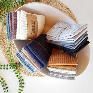 May include: A white bowl filled with stacks of waffle weave kitchen towels in various colors, including gray, blue, brown, and beige. The towels are tied with twine and have a label that reads "Flora et Lin".