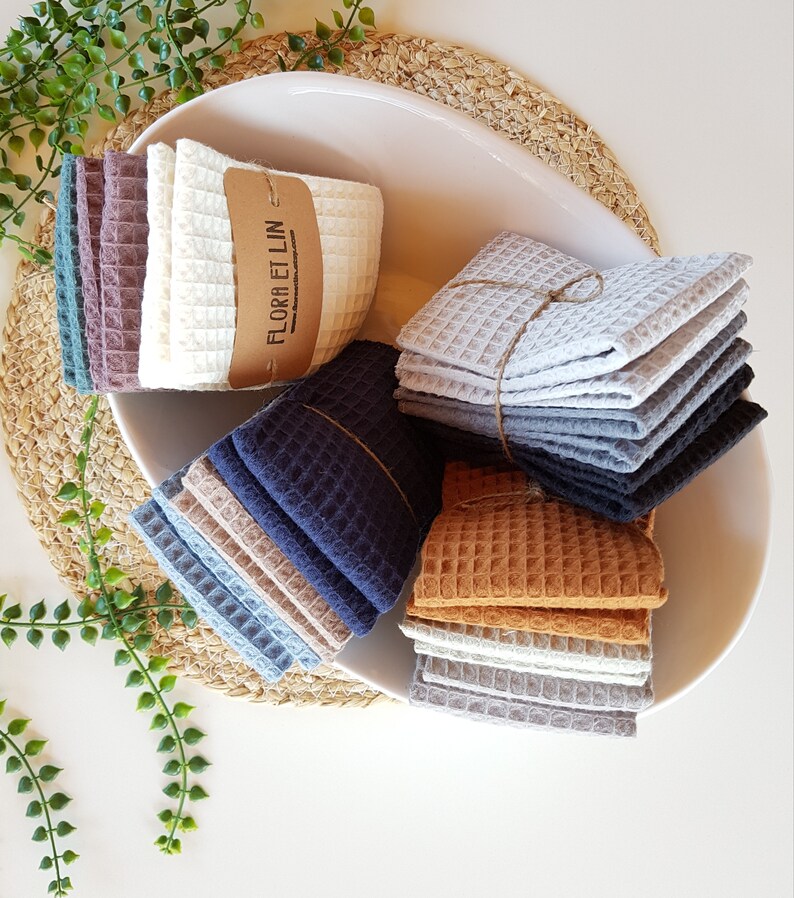 May include: A white bowl filled with stacks of waffle weave kitchen towels in various colors, including gray, blue, brown, and beige. The towels are tied with twine and have a label that reads "Flora et Lin".