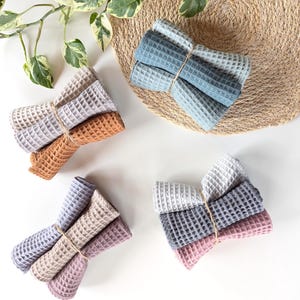 Peut inclure: Quatre ensembles de serviettes gaufrées de différentes couleurs, dont bleu, gris, beige et rose, attachées avec de la ficelle. Les serviettes sont disposées sur une surface blanche avec un dessous de plat tissé et de la verdure en arrière-plan.