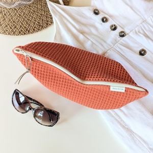 May include: A rust-coloured waffle-textured zippered pouch with a white zip and a small tag reading Flora Et Lin, accompanied by brown tortoiseshell sunglasses, a white button-down garment, and a woven straw hat brim in the background.