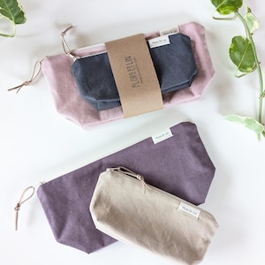 May include: A collection of fabric pouches in various sizes and colors. The set includes a large dusty rose pouch, a smaller charcoal gray pouch, and a medium-sized purple pouch. A tan pouch is also included. Each pouch has a zipper closure and a small tag that reads "Flora Et Lin".
