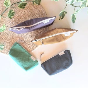 Peut inclure: Quatre pochettes en tissu de couleurs violet, beige, vert d'eau et gris anthracite. Chaque pochette a une fermeture éclair blanche et une petite étiquette blanche. Les pochettes sont disposées sur un tapis tissé et une surface blanche.