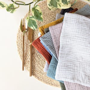 May include: A stack of colourful, textured cotton napkins in various shades of blue, purple, orange, and brown. The napkins are arranged on a woven straw placemat.