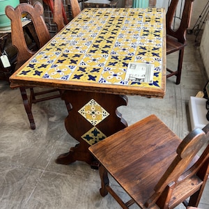 May include: A rectangular wooden table with a tiled top featuring a yellow, blue, and white floral pattern. The table has a dark brown wooden base and is surrounded by matching wooden chairs. The table's dimensions are approximately 60 inches long and 30 inches wide.