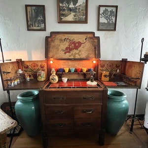May include: A vintage wooden bar cabinet with a painted scene of a bull on the front. The cabinet has four drawers and is flanked by two teal ceramic vases. The cabinet is decorated with colorful tiles and has a shelf with small colorful cups.