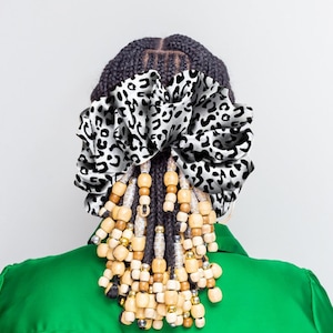 May include: A black and white leopard print fabric bow tied around a woman's braided hair with wooden and gold beads hanging down.