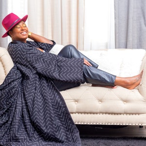 May include: A woman wearing a black and white chevron patterned robe and black leather pants sits on a cream-colored couch with a burgundy fedora on her head.