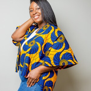 May include: A woman wearing a yellow and blue patterned kimono with a high low hemline. The kimono has a floral design with blue and yellow swirls. The woman is wearing denim shorts.