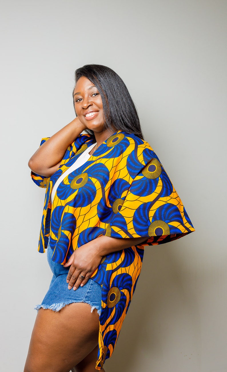 May include: A woman wearing a yellow and blue patterned kimono with a high low hemline. The kimono has a floral design with blue and yellow swirls. The woman is wearing denim shorts.