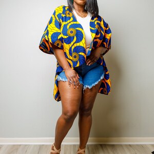 May include: A woman wearing a yellow and blue patterned kimono with a white top and denim shorts. She is standing on a wood floor and wearing tan sandals.