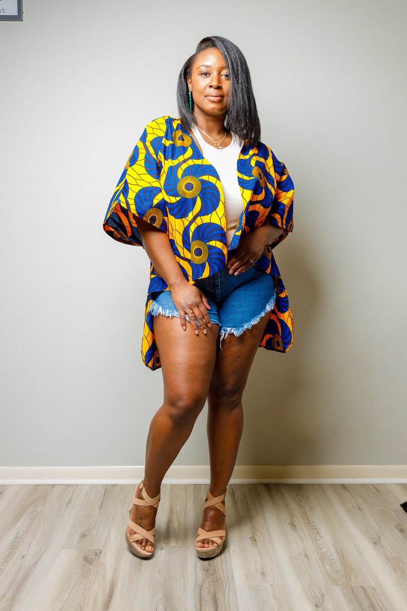 May include: A woman wearing a yellow and blue patterned kimono with a white top and denim shorts. She is standing on a wood floor and wearing tan sandals.