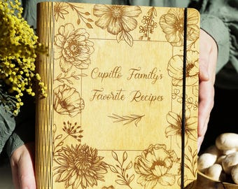 May include: A light-colored wooden recipe book with a black elastic closure. The cover features laser-engraved floral illustrations and the text "Cupillo Family's Favorite Recipes" centered within a rectangular border of various flower designs.