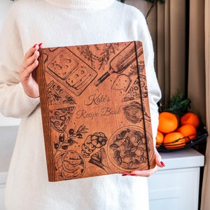 May include: A wooden recipe book with a brown cover and a design of various foods, including pizza, cheese, and olives. The text "Kate's Recipe Book" is engraved on the cover.