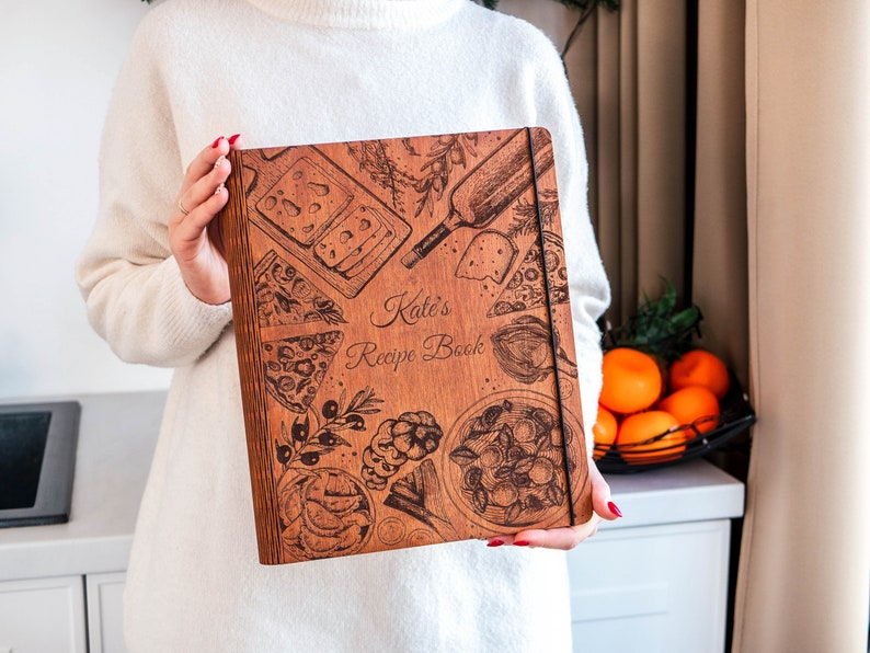 May include: A wooden recipe book with a brown cover and a design of various foods, including pizza, cheese, and olives. The text "Kate's Recipe Book" is engraved on the cover.