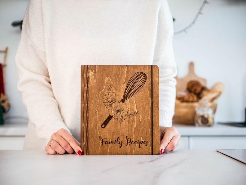 May include: A wooden recipe book with a floral design and a whisk engraved on the cover. The book is labeled "Family Recipes".
