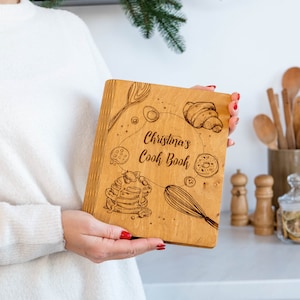 May include: A wooden cookbook with a personalized inscription that reads "Christina's Cookbook". The cover features a design of various baked goods, including croissants, donuts, and pancakes.