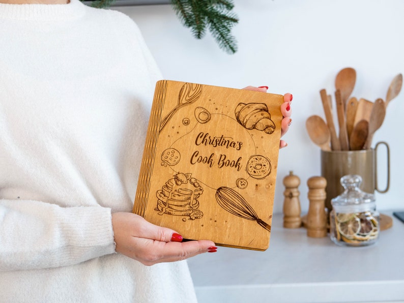 May include: A wooden cookbook with a personalized inscription that reads "Christina's Cookbook". The cover features a design of various baked goods, including croissants, donuts, and pancakes.