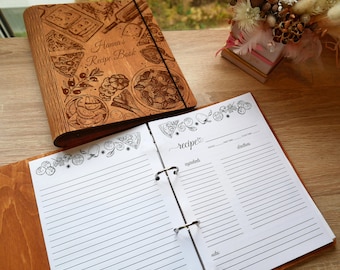 May include: A wooden recipe book with a cover featuring a pizza design and the text "Hanna's Recipe Book". The book is open to a blank page with lines for writing recipes. The page has a decorative border of pizza slices, tomatoes, and herbs.