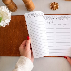 May include: An open recipe book with a brown leather cover. The pages are lined and have a decorative border of food illustrations. The book has sections for servings, prep time, cook time, ingredients, and instructions.