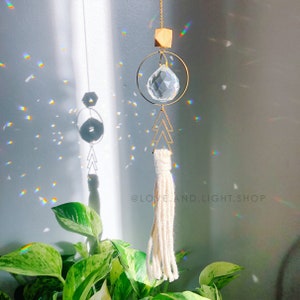 May include: A gold crystal suncatcher with a clear crystal ball, a gold geometric tree design, and a white tassel hanging from the bottom. The suncatcher is hanging from a window and casting rainbows on the wall. The text '@LOVELAND.LIGHT.SHOP' is visible in the image.