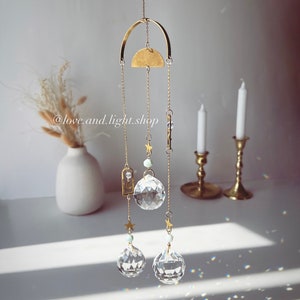 May include: A gold metal suncatcher with three crystal balls hanging from it. The suncatcher is decorated with small stars and a crescent moon. The suncatcher is hanging from a white ceiling.