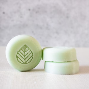 May include: Three round, pale green soap bars with a leaf design. The soap bars are stacked on top of each other.