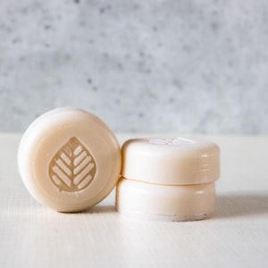 May include: Three round, off-white bars of soap. One bar stands upright on its edge, displaying a debossed leaf design on its front surface. The other two bars are stacked horizontally next to it, featuring a smooth, plain surface.