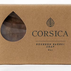 May include: A brown cardboard box with a teardrop-shaped window revealing a bar of soap. The box has the word "CORSICA" printed in black, along with "BOURBON BARREL SOAP" and "6 oz". A leaf design is above the word CORSICA.