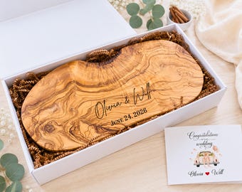 May include: An oval olive wood serving board with natural live edges and prominent wood grain, engraved with the text Olivia & Will and JUNE 24, 2026, nestled in a white gift box with brown paper filler and a wedding greeting card.