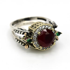 Könnte beinhalten: Ein silberner Ring mit einem großen, runden, tiefroten Edelstein in der Mitte, umgeben von kleineren weißen Steinen. Der Ring hat aufwendige Details mit goldenen Akzenten und kleineren grünen und roten Steinen.