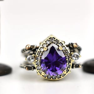 Könnte beinhalten: Ein silberner Ring mit einem birnenförmigen, tiefvioletten Edelstein in der Mitte, umgeben von kleinen weißen Steinen und einem goldfarbenen Rand. Der Ring hat ein mehrschichtiges Design mit zusätzlichen kleinen weißen Steinen. Zwei bronzefarbene Dekorfiguren im Hintergrund.