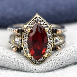 May include: A silver and gold ring set with a large, marquise-cut, deep red gemstone as the focal point. The ring is surrounded by smaller white gemstones and gold accents. The ring sits on a white and blue fabric.