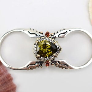 Könnte beinhalten: Ein silberner Ring mit einem birnenförmigen grünen Edelstein, der von kleineren klaren Steinen umgeben ist. Der Ring hat ein dekoratives Design mit Schnörkeln und einem Doppelband.