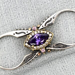 Könnte beinhalten: Silberfarbene Brosche mit einem ovalen, tiefvioletten Edelstein in der Mitte, umgeben von kleinen weißen und goldenen Akzenten. Die Brosche hat ein einzigartiges, offenes Design mit geschwungenen Schlaufen und kleinen rosa Edelsteinen.