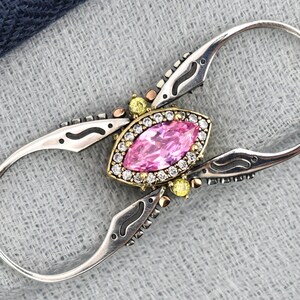 Könnte beinhalten: Ein silberfarbener Ring mit einem ovalen, rosa Edelstein in der Mitte, umgeben von kleineren weißen und gelben Steinen. Der Ring hat ein aufwendiges Design mit gebogenen, länglichen Enden. Der Hintergrund ist ein strukturierter grauer Stoff mit einem dunkelblauen Abschnitt.