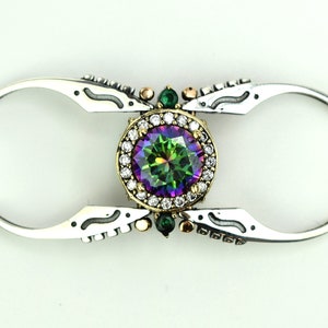 Könnte beinhalten: Ein silberner Ring mit einem großen, runden, schillernden grünen und violetten Edelstein, der von einem Heiligenschein aus kleineren weißen Edelsteinen umgeben ist. Der Ring ist an beiden Seiten offen und hat ein dekoratives Design.